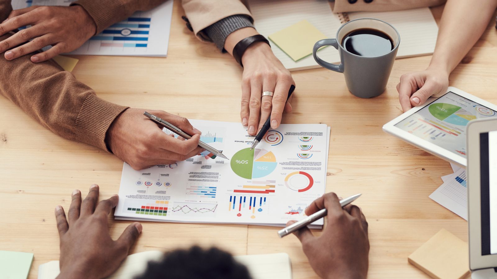 Hands pointing to printed charts on a table beside a tablet and a cup of coffee.